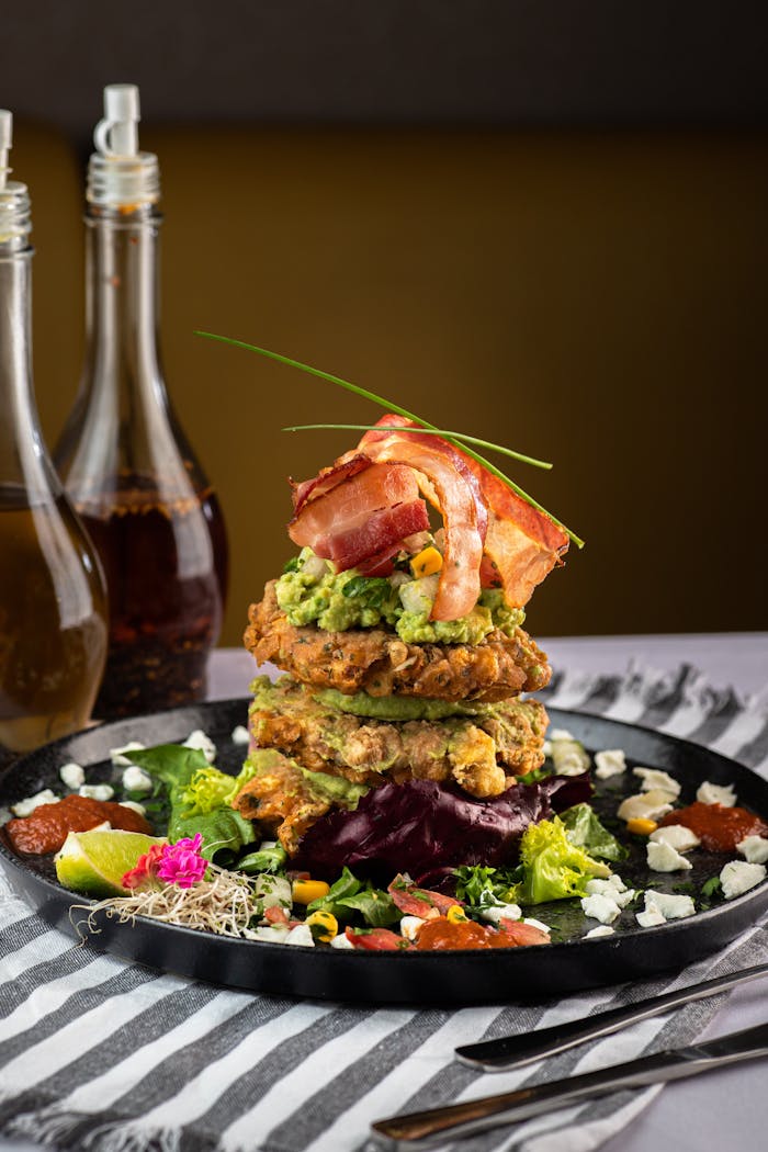 Enticing gourmet pancake stack with bacon, avocado, and vibrant salad on a black plate.